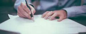 A man signing some paper on a desk with a blue suit